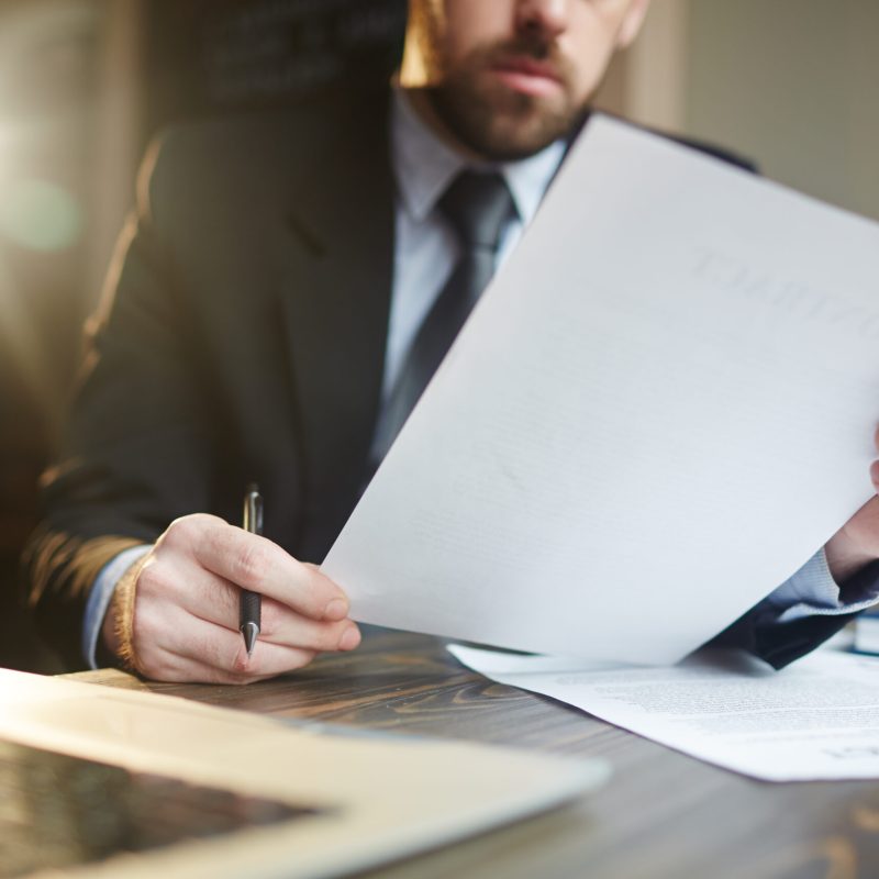 Portrait of modern bearded businessman holding papers in hands, reading and analyzing contract documentation at desk with laptop